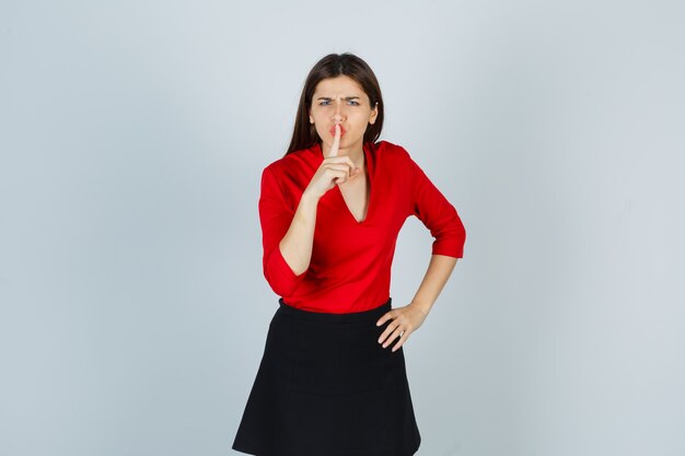 Young lady showing silence gesture in red blouse, black skirt and looking angry