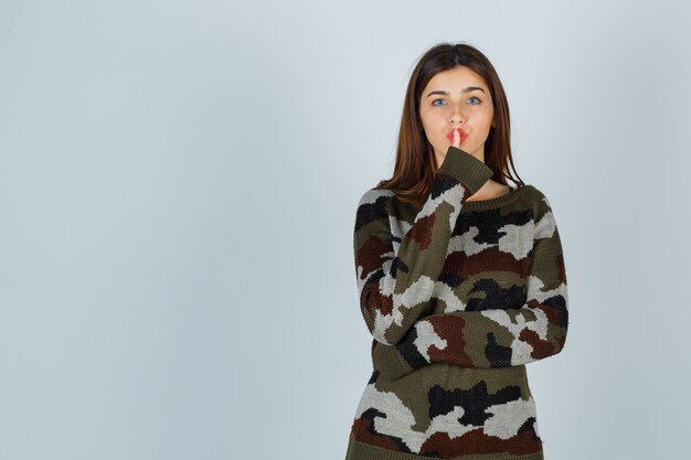 Young lady showing silence gesture in sweater, skirt and looking careful