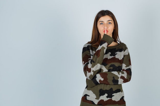 Young lady showing silence gesture in sweater, skirt and looking careful