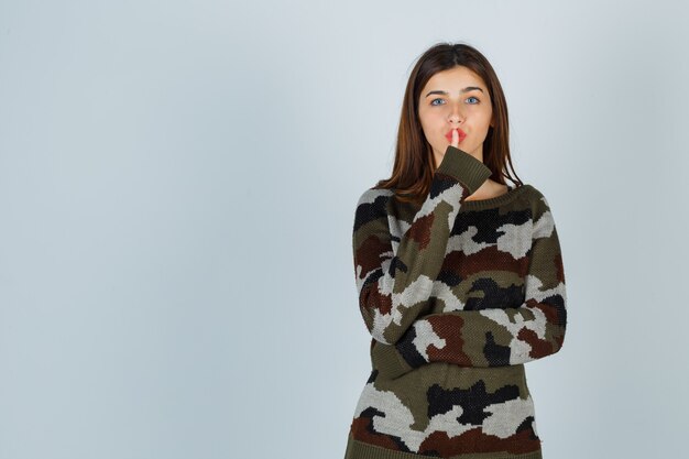 Молодая дама показывает жест молчания в свитере, юбке и осторожно смотрит