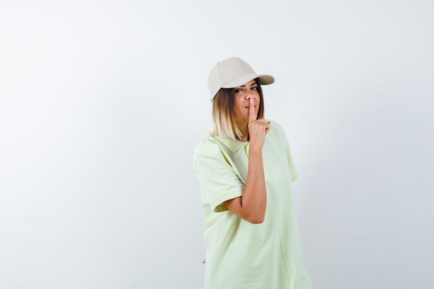 Tシャツ、キャップ、自信を持って、正面図で沈黙のジェスチャーを示す若い女性。