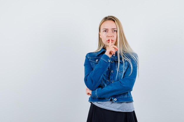 Giovane donna che mostra gesto di silenzio in maglietta, giacca di jeans, gonna e sembra sicura and