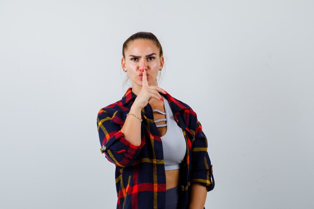 Young lady showing silence gesture in top, plaid shirt and looking serious , front view.