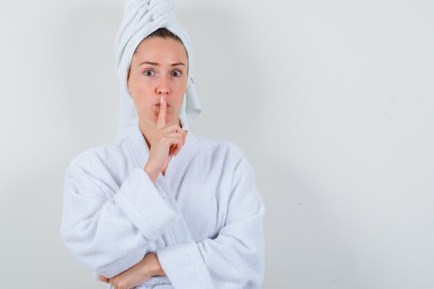 Young lady showing silence gesture in white bathrobe, towel and looking careful , front view.