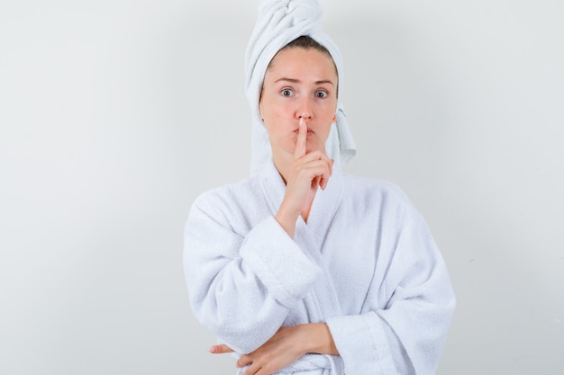 Young lady showing silence gesture in white bathrobe, towel and looking sensible , front view.