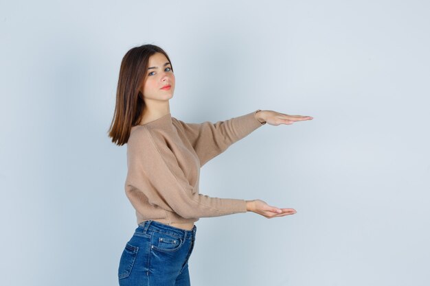 Young lady showing size sign in beige sweater, jeans and looking pleased. front view.