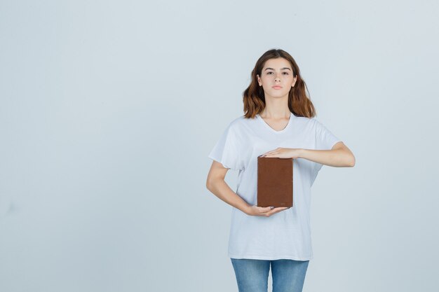 Tシャツ、ジーンズ、自信を持って、正面図で本を保持しながらサイズサインを示す若い女性。