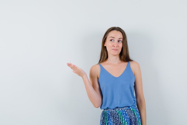 Young lady showing something with hand in singlet, skirt and looking curious. front view.