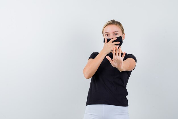 停止ジェスチャーを示している若い女性、黒いTシャツ、マスク、怖い顔、正面図で手を口に保ちます。
