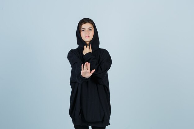 Young lady showing stop gesture in oversized hoodie, pants and looking serious , front view.