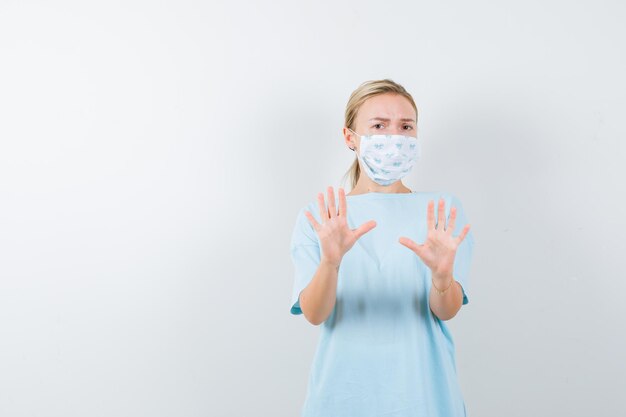 Young lady showing stop gesture in t-shirt, mask and looking scared isolated