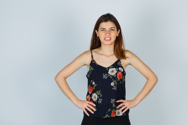 Young lady showing teeth, holding hands on waist in floral top and looking gloomy