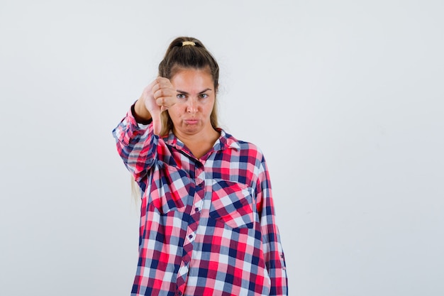 Young lady showing thumb down in checked shirt and looking displeased , front view.