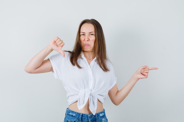 Young lady showing thumb down while pointing aside in white blouse and looking negative. front view.