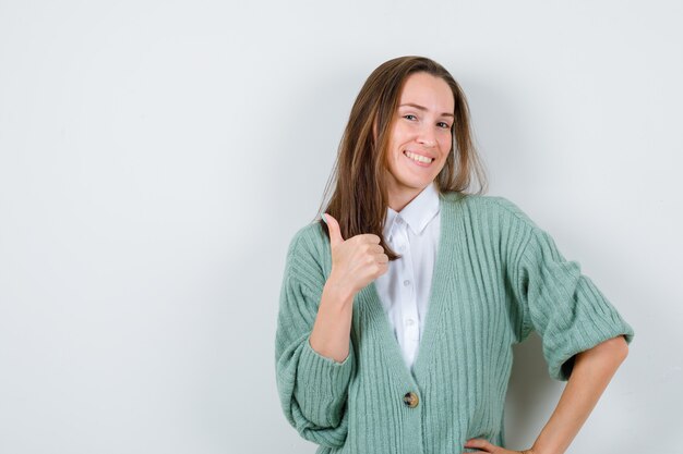 Young lady showing thumb up in shirt, cardigan and looking cheery , front view.