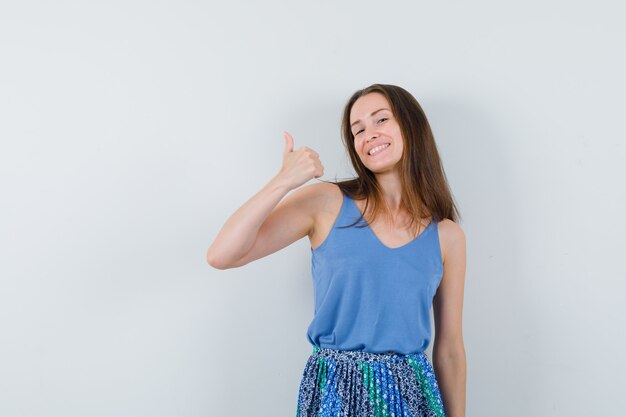 Young lady showing thumb up in singlet, skirt and looking happy , front view.