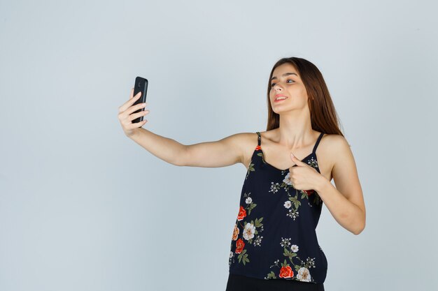 Young lady showing thumb up on video call in blouse and looking glad. front view.