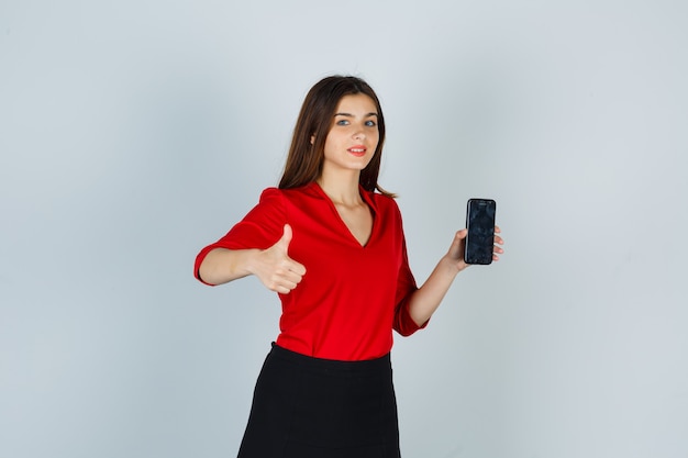 Девушка показывает палец вверх, держа мобильный телефон в красной блузке