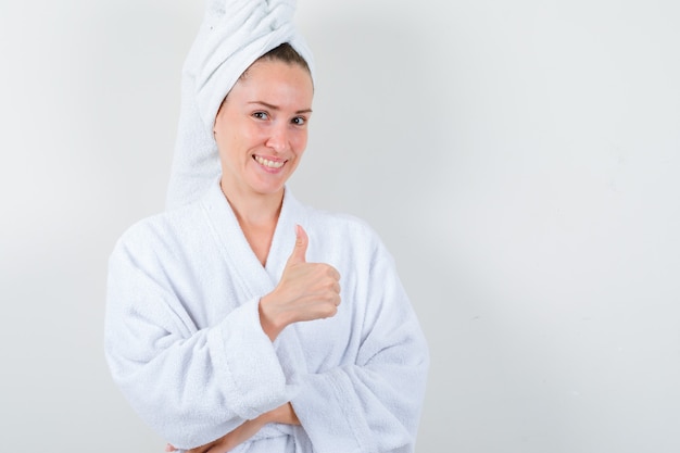 Девушка показывает палец вверх в белом халате, полотенце и выглядит весело. передний план.