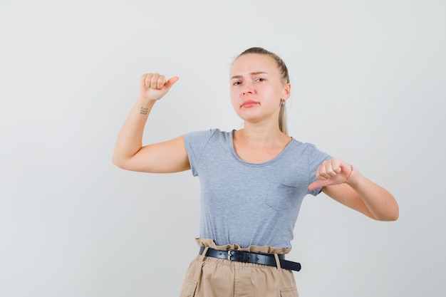 Young lady showing thumbs up and down in t-shirt and pants and looking indecisive