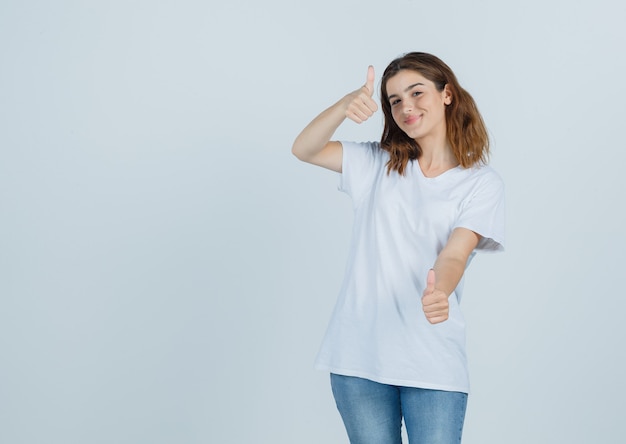 Young lady showing thumbs up in t-shirt, jeans and looking joyful , front view.