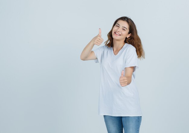 Young lady showing thumbs up in t-shirt, jeans and looking joyful , front view.