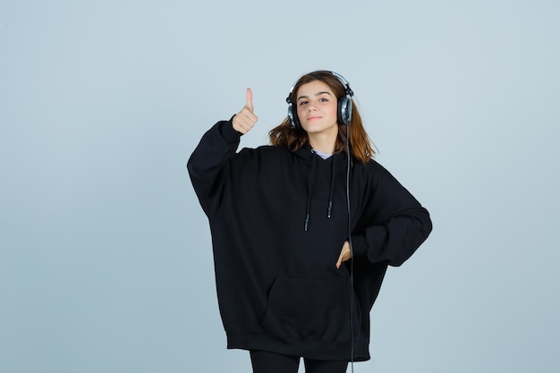 Foto gratuita giovane donna che mostra i pollici in su mentre tiene la mano sulla vita in felpa con cappuccio oversize, pantaloni e sembra beata, vista frontale.