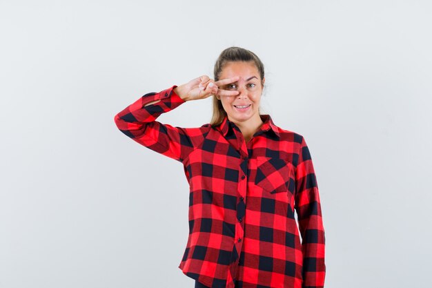 Young lady showing V-sign on eye in checked shirt and looking confident