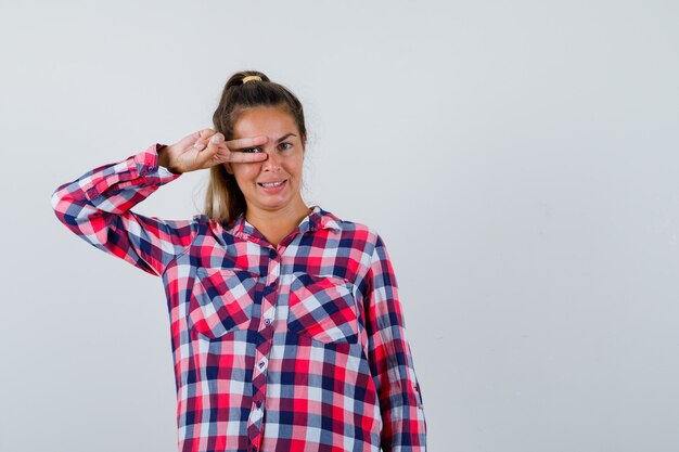Young lady showing V-sign on eye in checked shirt and looking happy , front view.