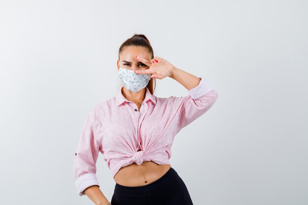 Young lady showing V-sign on eye in shirt, pants, mask and looking merry , front view.