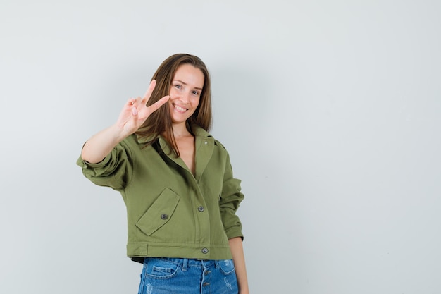 Young lady showing v-sign in green jacket shorts and looking confident  