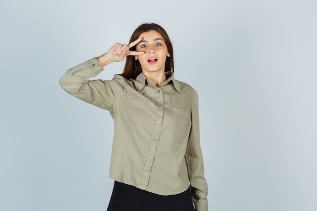 Young lady showing V-sign near eye in shirt, skirt and looking wondered , front view.