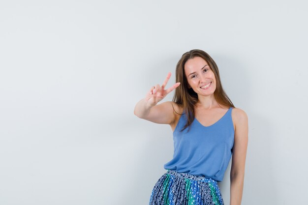 Young lady showing V-sign in singlet, skirt and looking happy. front view.