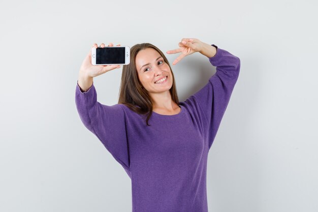 Young lady showing v-sign while taking photo in violet shirt and looking merry , front view.