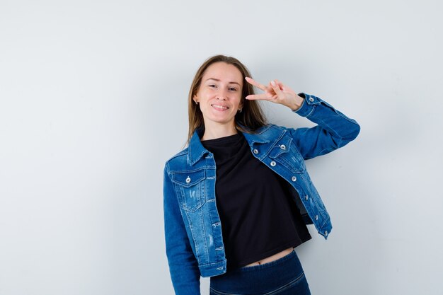 Young lady showing victory gesture in blouse, jacket and looking confident , front view.