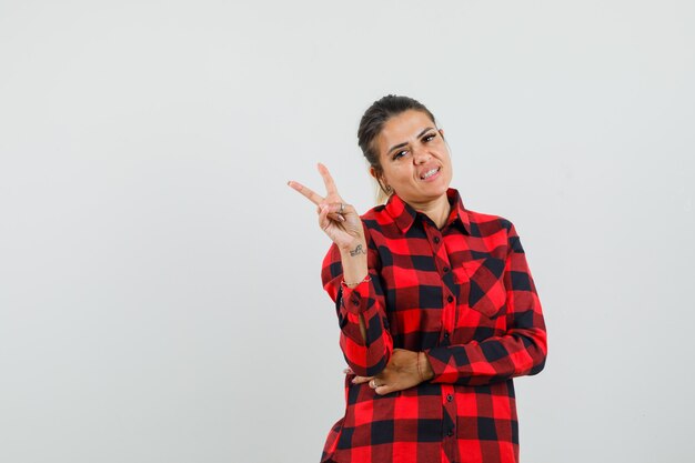 Young lady showing victory gesture in checked shirt and looking confident