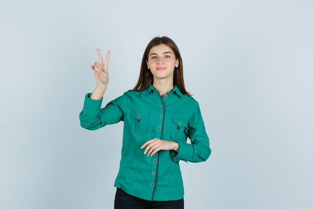 Young lady showing victory gesture in green shirt and looking cheerful , front view.