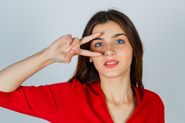 Young lady showing victory sign on eye in red blouse and looking lucky