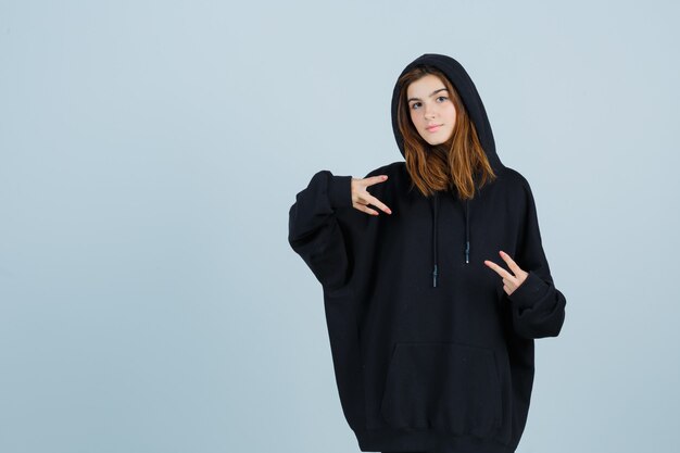 Young lady showing victory sign in oversized hoodie, pants and looking confident. front view.