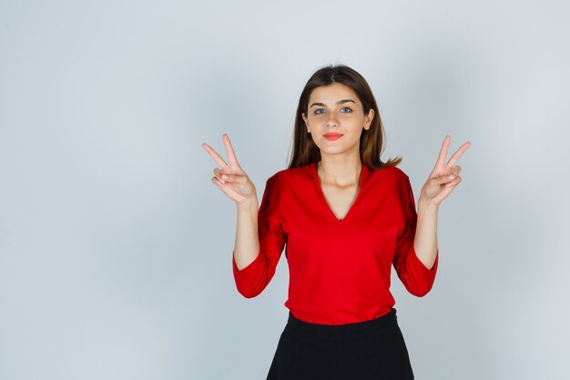 Young lady showing victory sign in red blouse, skirt and looking cheery  