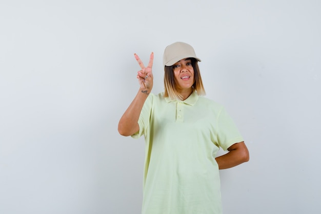 Young lady showing victory sign in t-shirt, cap and looking blissful , front view.