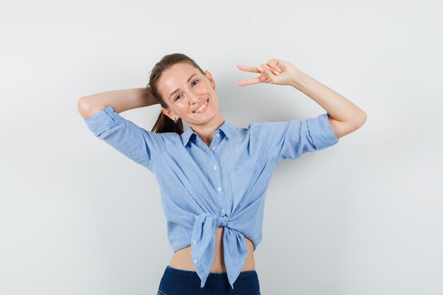 Young lady showing victory sign while posing in blue shirt, pants and looking joyful.