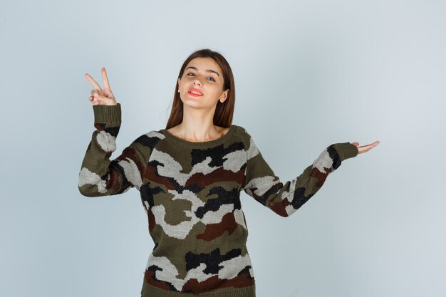 セーターで何かを示すふりをしながら勝利のサインを示す若い女性
