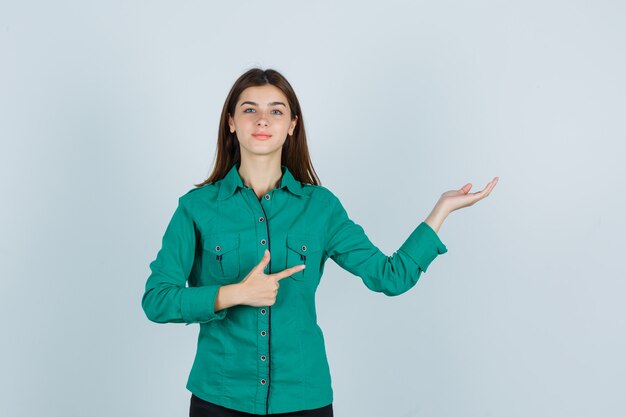 Молодая леди показывает приветливый жест, указывая в сторону в зеленой рубашке и выглядит уверенно, вид спереди.