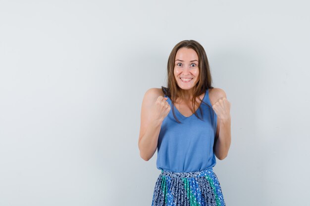 Young lady showing winner gesture in blue blouse,skirt and looking merry. front view.