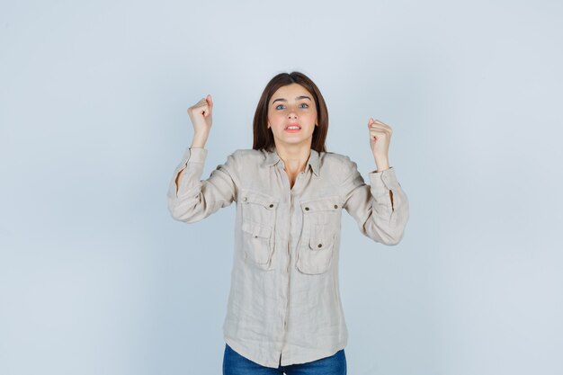 Young lady showing winner gesture in casual, jeans and looking victorious. front view.