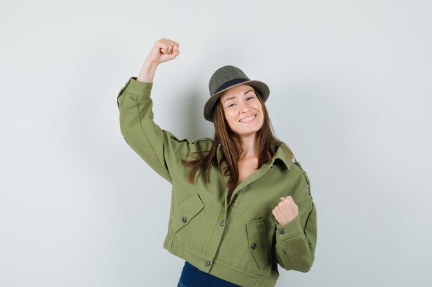 ジャケットパンツの帽子で勝者のジェスチャーを示し、至福に見える若い女性