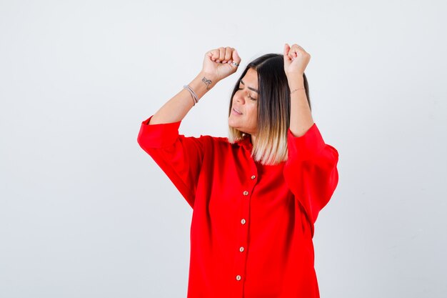 Young lady showing winner gesture in red oversize shirt and looking lucky , front view.