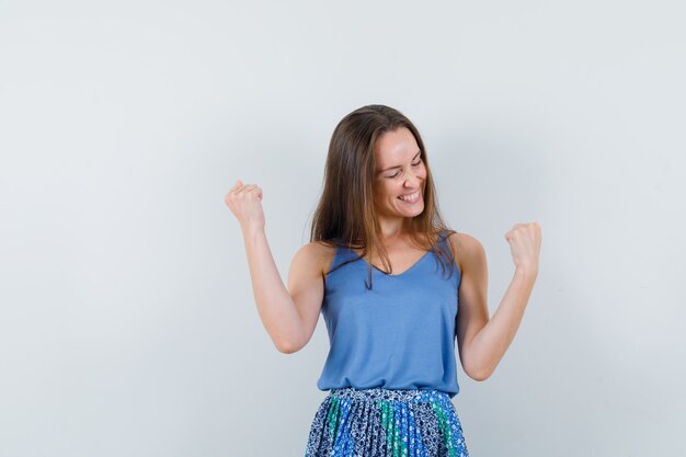 Young lady showing winner gesture in singlet, skirt and looking merry , front view.