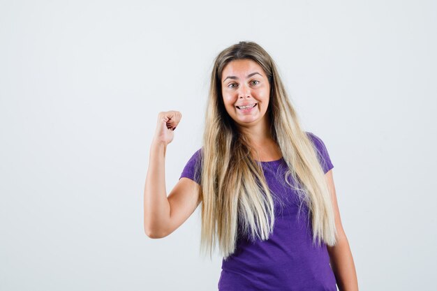 紫のTシャツで勝者のジェスチャーを示し、幸せそうに見える若い女性、正面図。